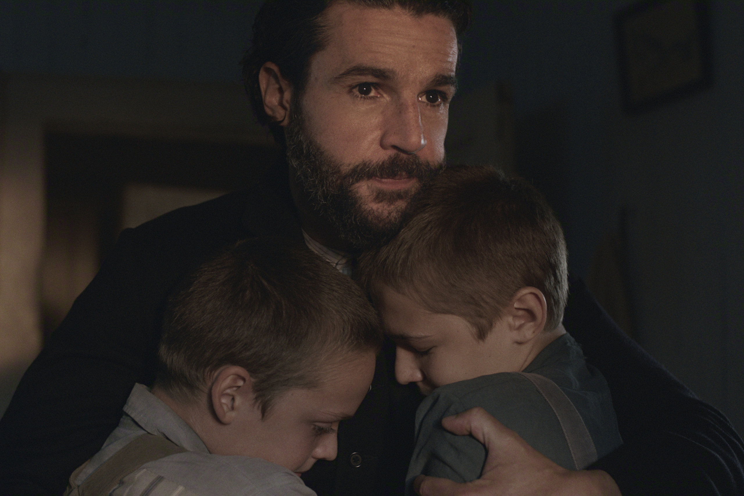 "A bearded man in a dimly lit room hugs two young boys close to his chest, providing comfort. All three have serious, emotional expressions, suggesting a moment of sadness or reassurance in a home setting."
